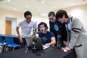 Students wearing Google shirts exclaim at a laptop sitting next to two Redhat water bottles.
