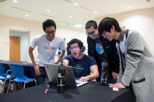 Students wearing Google shirts exclaim at a laptop sitting next to two Redhat water bottles.