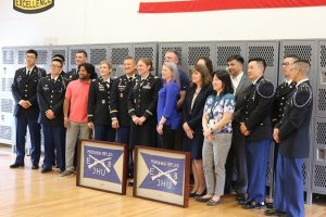 ROTC members, including Jenna Ferrell, pose in front of lockers. In front of them are two framed blue flags with "PERISHING RIFLES E 8 JHU" stitched on them around with two crossed rifles.