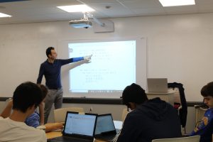 Mohammad Ali Darvish gestures to code being projected on a screen. Students observe with their laptops open.