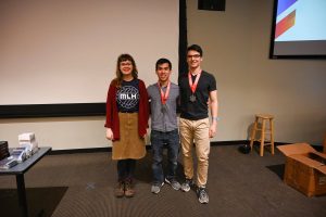 Katie O'Leary (a Major League Hacking Representative), Travis Chan, and Thomas Keady pose together at the front of Hodson 110. Chan and Keady have medals around their necks.