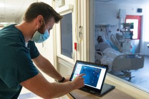Wearing a mask, Axel Krieger points at a tablet showing a patient's health status outside of the patient's hospital room.