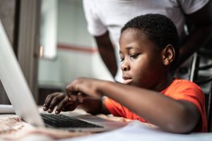 A young boy types on a laptop. A parent watches over his shoulder.