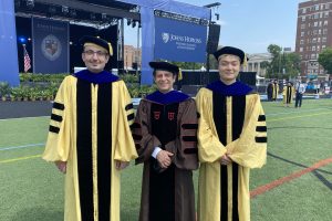 Ehsan Azimi, Peter Kazanzies, and Long Qian pose on Homewood field wearing graduation regalia in front of a sign that reads Johns Hopkins Whiting School of Engineering. Azimi and Qian wear yellow robes. Kazanzides wears a brown robe.