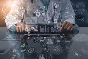 A doctor wearing a white coat and stethoscope places their fingers on a blank tablet laid on a flat surface. The surface is overlaid with hexagons, medical clip art, and graphs, which are also hovering mid-air in front of the doctor. The doctor's head is cropped.