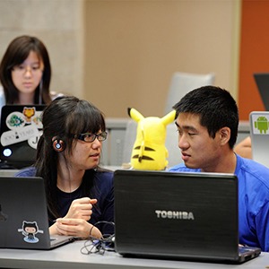 Students at laptops discuss in class. A toy Pikachu sits on top of one laptop.
