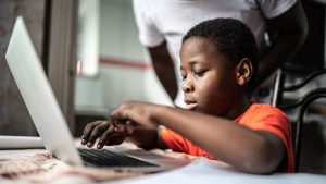 A young boy types on a laptop with his parent supervising.