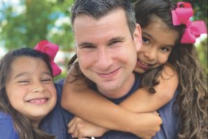 Aaron Baughman with his two daughters. They wear pink ribbons in their hair and embrace him.