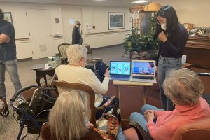A student wearing a mask stands next to two laptops on a folding table in a nursing home. Three older women watch her demonstration.
