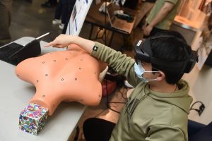 A student wearing a mask and augmented reality glasses gestures to a plastic torso with white dots on it. In place of a head the torso has a colorful box.