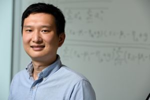Headshot of Yinzhi Cao standing in front of a whiteboard with mathematical equations written on it.