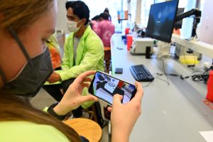 A student wearing a mask demonstrates the Quest2Learn augmented reality application on their smartphone. The smartphone displays a lab setup superimposed on the empty desk in front of the student.
