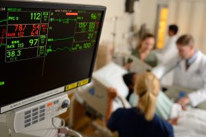 An image of a hospital room with the focus on the patient's health statistics. Out of focus, the patient is lying in bed surrounded by doctors and nurses.