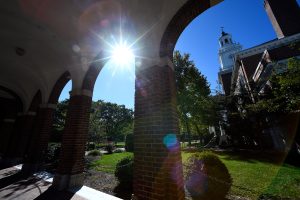 A view of Gilman Hall through the arches on its left side with the sun shining brightly in the blue sky.