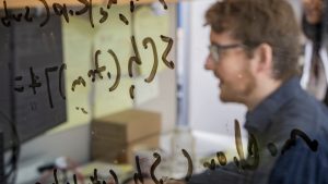 A student looking at a computer screen, viewed through glass with equations marked on it in marker.