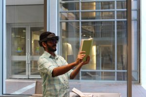A student solves a mixed reality escape room. He wears a HoloLens and holds a book up in front of him, making gestures with his right hand.