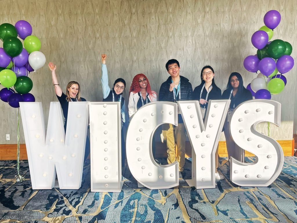 The JHU Women in Cybersecurity chapter standing behind large WICYS letters. There are white, purple, and green balloons on either side of the group.