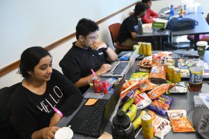 Students code on their laptops at a table overflowing with snacks.