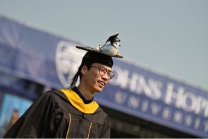 A master's graduate in commencement hood and robes with a stuffed blue jay on his graduation cap.