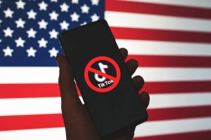 A hand holds a phone with a red circle and line around and through the TikTok logo. An American flag is in the background.
