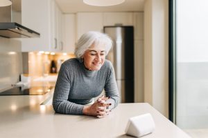 An older woman leans against a kitchen counter and smiles at a smart speaker.