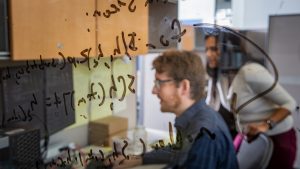 View of students looking at a computer through glass covered in mathematical equations.