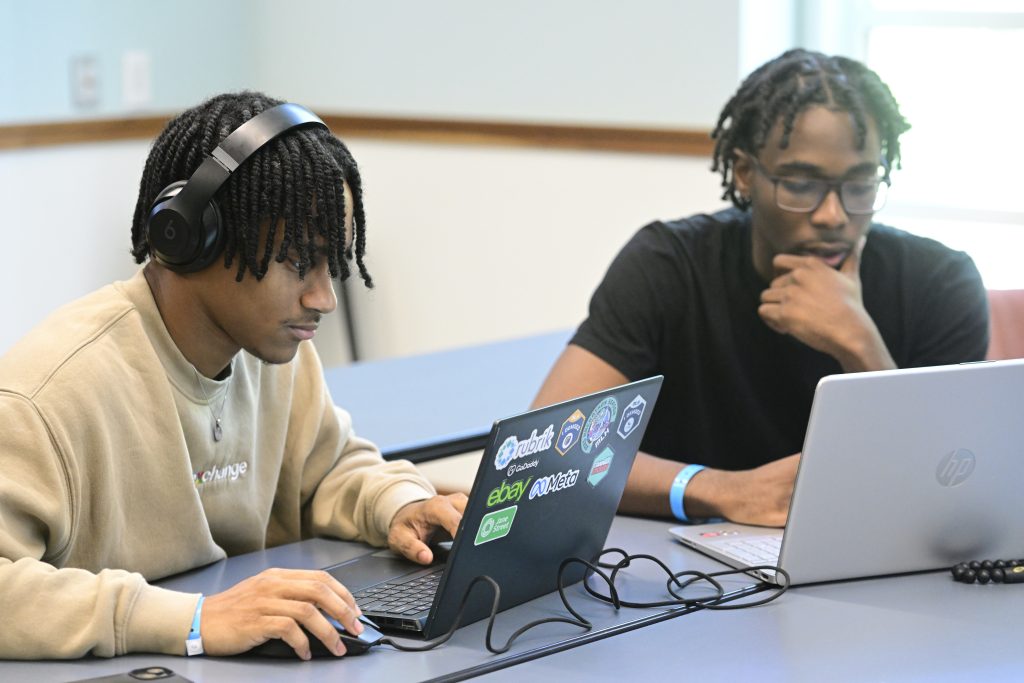 Two students with dreadlocks hunch over their laptops in concentration.