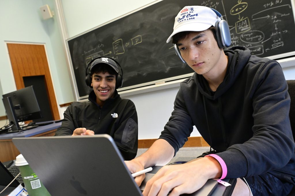 A student with headphones on types on a laptop as another smiling student looks on.