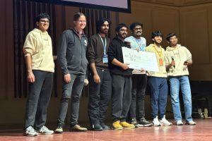Joy Bhalla (center), Bontalakoti Venkata Harshavardhan (immediate left), and Arnab Maity (immediate right) pose with a large check with "1st Place" written on it alongside NexHacks organizers and sponsors.