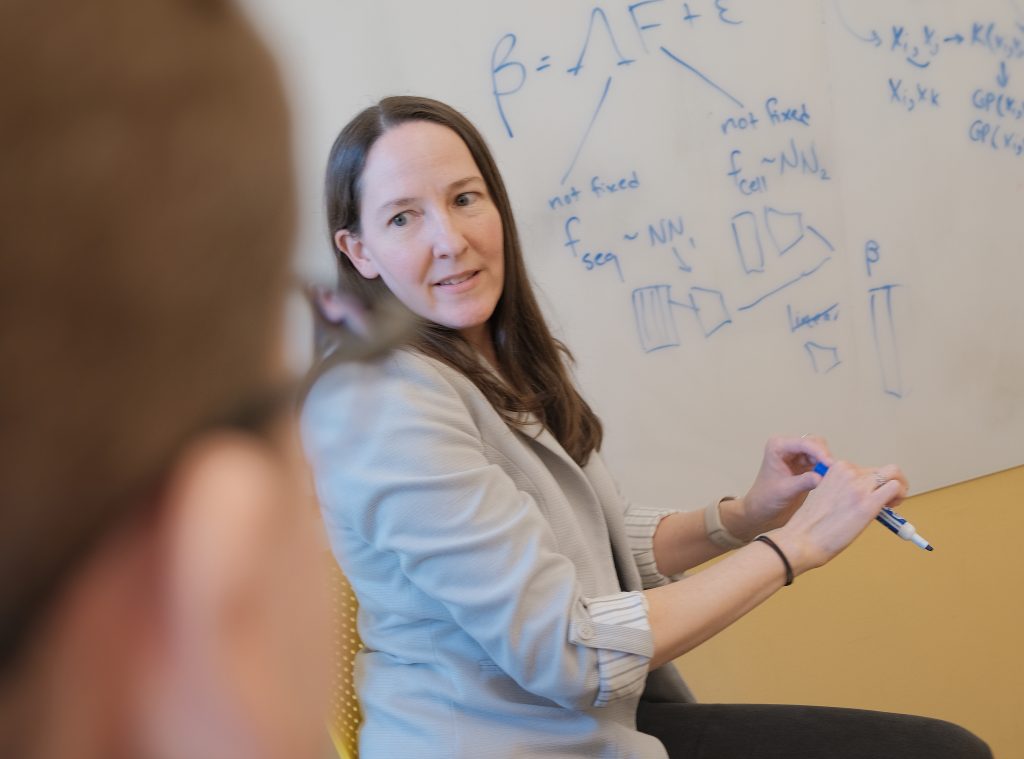 Alexis Battle in front of a whiteboard with a complex formula on it, holding a dry-erase marker and speaking to someone in the foreground.