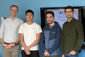 Ben Langmead, Steven Tan, Mohsen Zakeri, and Sina Majidian stand in front of a TV screen with a picture of a Best Paper Award on it.