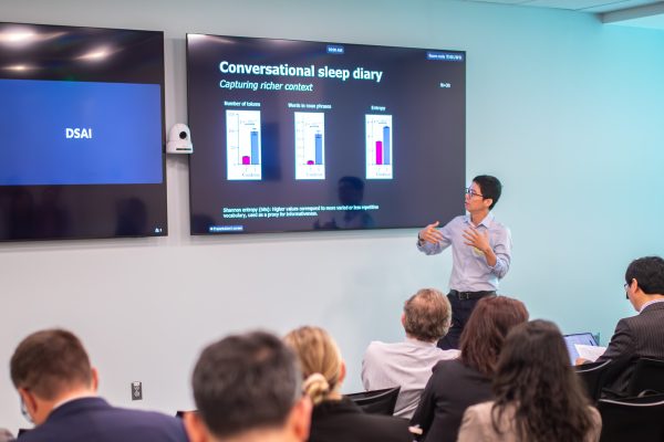 Chien-Ming Huang presents to an audience in front of a TV screen with three bar graphs that reads "Conversational sleep diary: Capturing richer context."